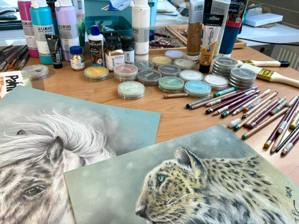 Arbeitstisch im Atelier mit Acrylfarben, Pastellkreiden, Buntstiften und zwei Tierporträts.